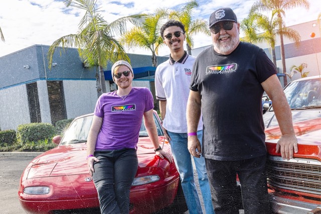 Three men, one older, two younger, standing in front of two red class cars