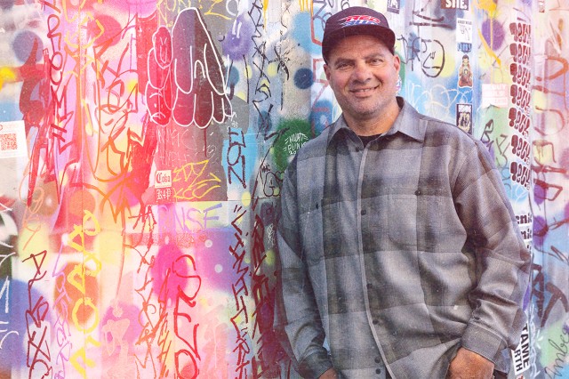 Man standing in front of a call covered in colorful graffiti, leaning his right shoulder against the wall. His hands are in his pockets; he wears a dark baseball cap and gray plaid, button-down long-sleeved shirt.