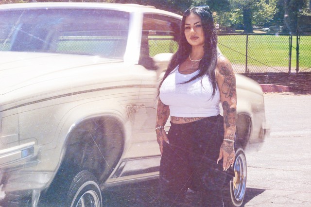 Latino woman in a white tank top and black capris standing next to a white lowrider car.
