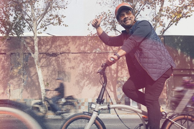 Man in black pants, blue short and baseball hat riding a bike and giving a thumbs up