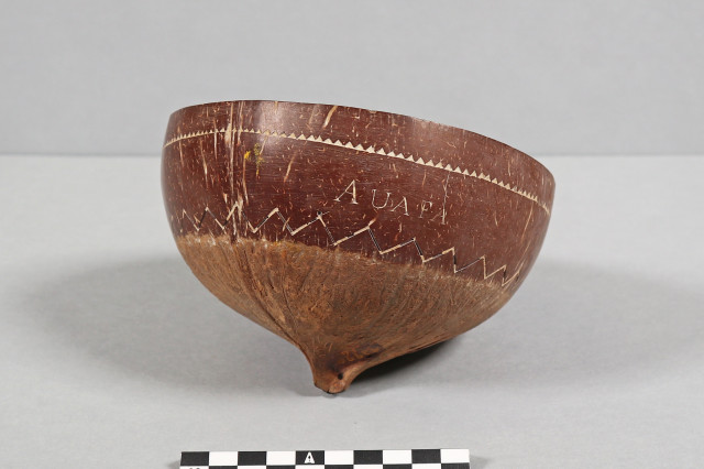 A wide-angle, eye-level studio photograph shows a deep, rounded bowl crafted from a polished coconut shell. The bowl is tilted slightly to the right, resting on a small, pointed nub at its base. It sits against a neutral, light-gray background. A black-and-white centimeter scale bar is positioned in the foreground for size reference.
