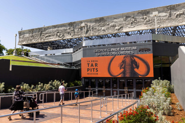 Visitors walking on a path towards the entrance of La Brea Tar Pits Museum, with an orange banner over the doorway