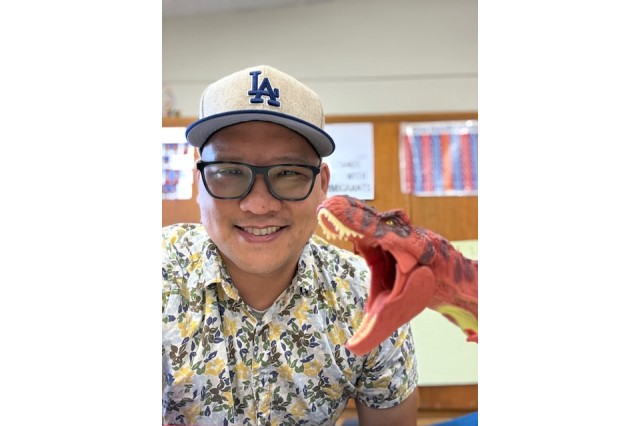 David Kwan wearing a white baseball cap with LA in blue letters, black glasses, and a floral patterned shirt, next to a red dinosaur figurine with its mouth open