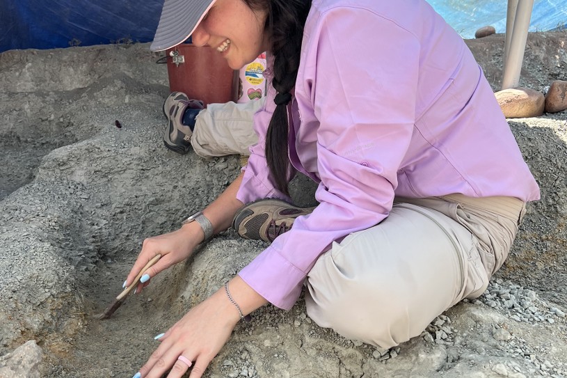 Bridget Altman wearing a hat and pink shirt, holding a brush while sitting and leaning over a carved out rectangle in the dirt