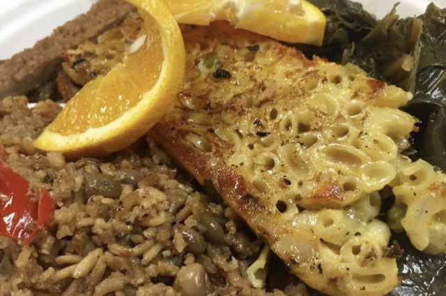 An overhead view of the Island Delight plate featuring a square of golden-brown baked macaroni pie, a portion of pigeon peas and rice, and sautéed greens, garnished with fresh orange slices and plantains.