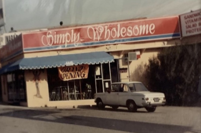 A historical black-and-white photo of the original Simply Wholesome storefront in 1984, featuring a "Grand Opening" banner hanging above the entrance.