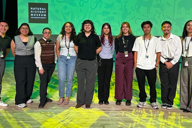 UNLAB participants standing in a row on stage with yellow lighting and a green image on a screen in the background