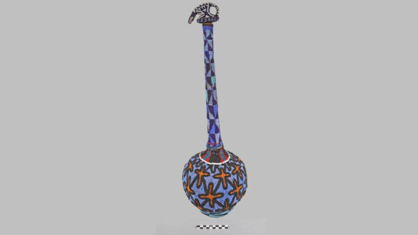 A full-length, vertical studio photograph features a tall, slender-necked gourd vessel completely covered in intricate glass beadwork. The object stands on a flat, neutral gray surface against a light gray background. It is topped with a decorative animal-shaped stopper. A black-and-white 10-centimeter scale bar is positioned at the bottom center.