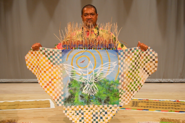 Jason Pereira holding up a checker-patterned woven mat with an attached painting of a white-outlined bird