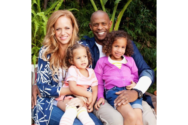 Family photo taken outdoors of a mixed race family of four. Two young girls sit in their parents laps.
