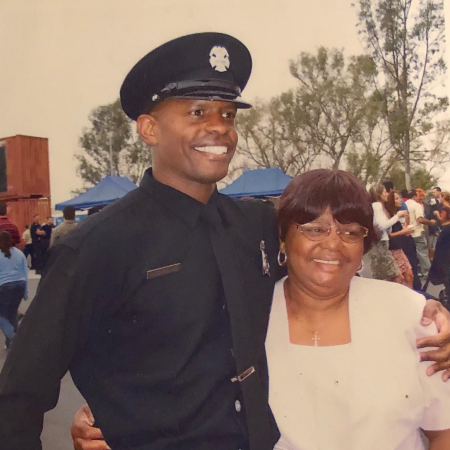 Black man in an LAFD Academy uniform posing outside, next to his mother on graduation day.