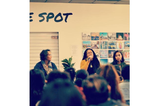 Three women on a panel in front of a seated crowd in a coffee shop; one woman talks into a microphone.